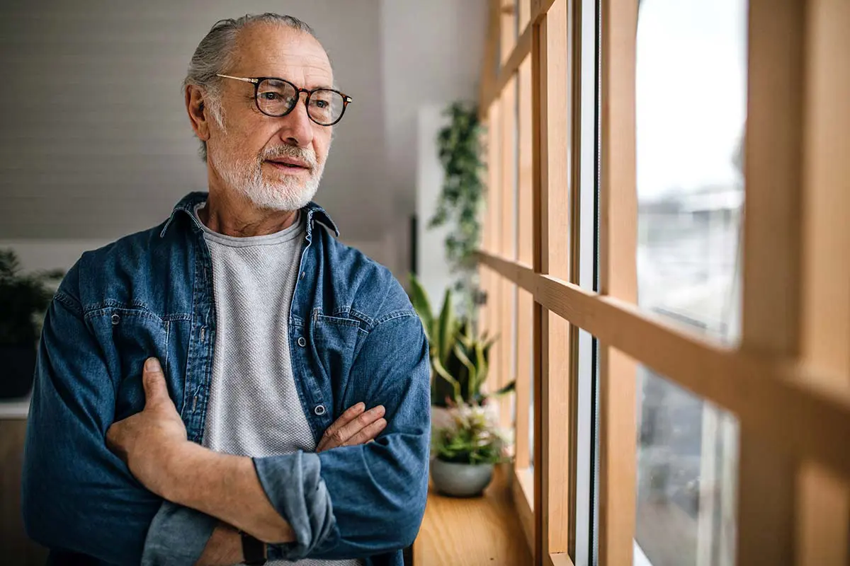 A senior man wearing a denim shirt looks out the window on a cloudy day.