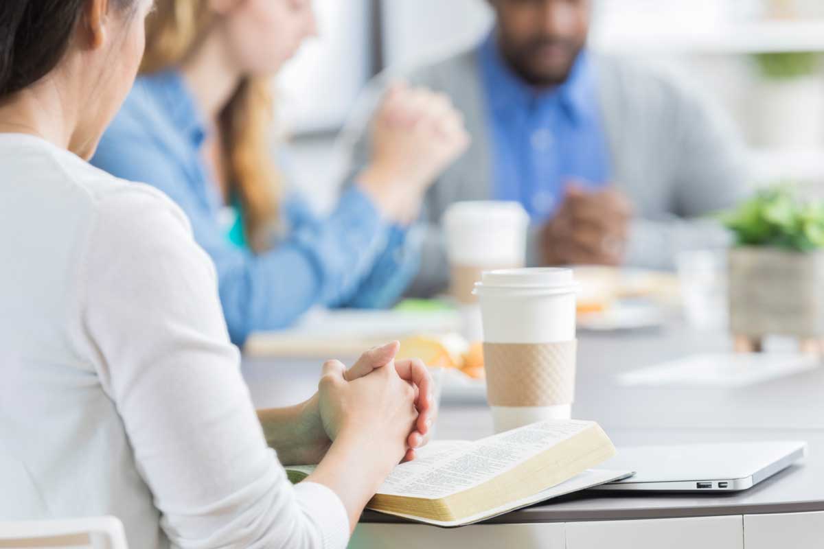 Church administrators pray with the Bible and a laptop on a desk during a church admin meeting.