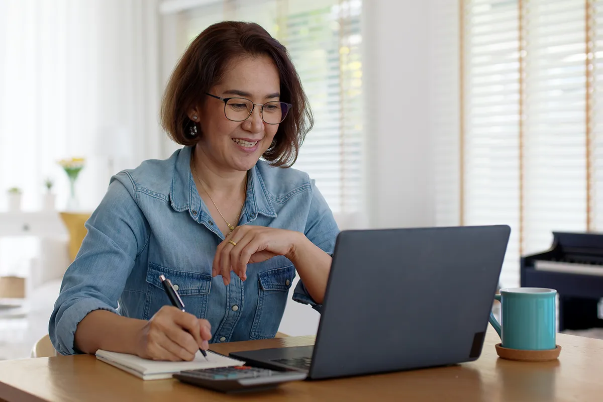 A smiling woman taking notes while watching an online course on her laptop.