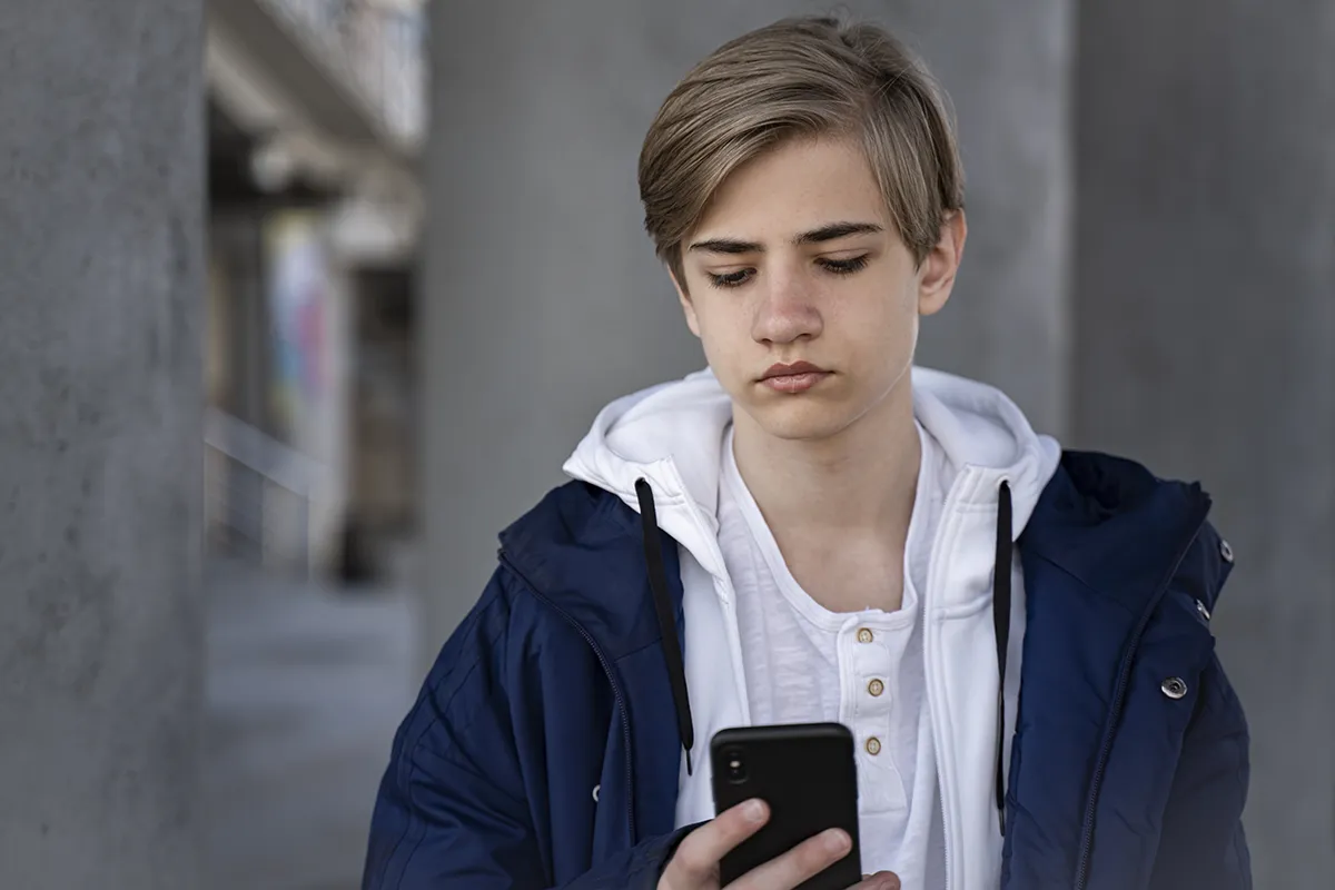 A concerned teen boy looking at his smartphone. Find out how teen boys are becoming victims of sextortion scams.